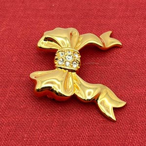 Op de afbeelding: Een gouden strik broche met heldere strass steentjes in het midden.