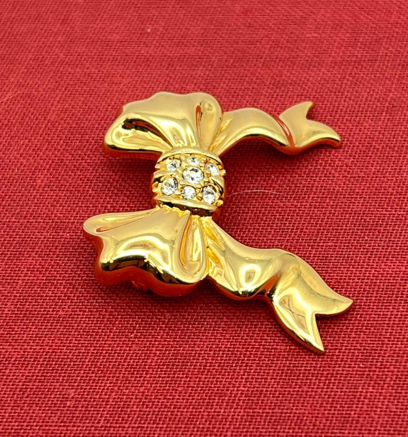 Op de afbeelding: Een gouden strik broche met heldere strass steentjes in het midden.