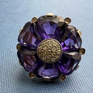 May include: A flower-shaped ring featuring six large, faceted, deep purple stones arranged as petals around a central circular cluster of small, clear, pavé-set crystals. The stones are held in place by a metallic setting with small prongs.