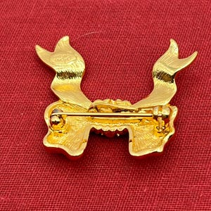 Op de afbeelding: Gouden broche in de vorm van een strik met een pin aan de achterkant. De broche heeft een getextureerd oppervlak en een klein opschrift aan de achterkant dat "SWAROVSKI" luidt.