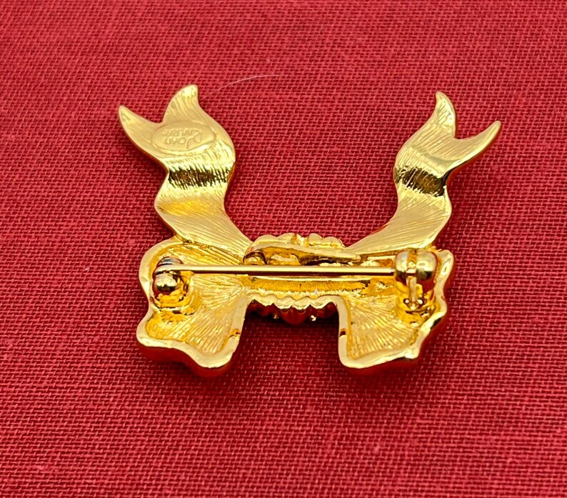Op de afbeelding: Gouden broche in de vorm van een strik met een pin aan de achterkant. De broche heeft een getextureerd oppervlak en een klein opschrift aan de achterkant dat "SWAROVSKI" luidt.