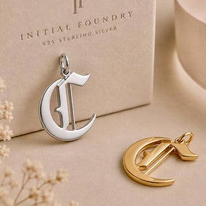 May include: A silver-colored letter C pendant and a gold-colored letter C pendant, both featuring an old English font style. The silver pendant is attached to a beige box with the text IF INITIAL FOUNDRY 925 STERLING SILVER printed on it.