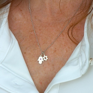 Puede incluir: Un collar de plata con un dije de mano de Hamsa y un dije de estrella de David. El dije de mano de Hamsa tiene una pequeña piedra rosa en el centro.