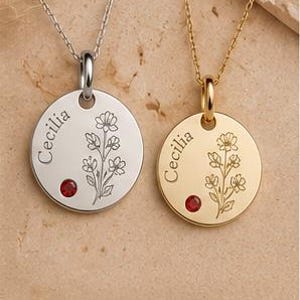 Könnte beinhalten: Zwei runde Kettenanhänger, einer in Silber und einer in Gold, jeweils mit dem eingravierten Namen Cecilia, einer floralen Strichzeichnung und einem kleinen roten Edelstein. Der Text Available in Sterling Silver & Gold steht unten.