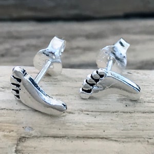 May include: A pair of silver stud earrings featuring a footprint design. The earrings are small and delicate, perfect for everyday wear.
