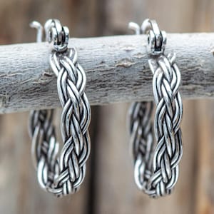 May include: A pair of silver hoop earrings with a braided design. The earrings are displayed on a wooden branch against a blurred background. The braided pattern gives the earrings a textured appearance.