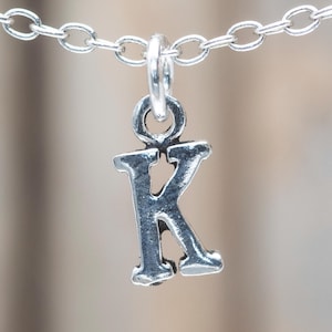 Op de afbeelding: Een zilverkleurige ketting met een kleine, gedetailleerde letter "K" hanger. De hanger hangt aan een fijne ketting, en de achtergrond is wazig, waardoor de aandacht op de sieraden wordt gevestigd.
