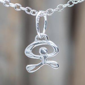 Op de afbeelding: Zilveren ketting met een hanger in de vorm van een gestileerde figuur in een yogahouding. De hanger hangt aan een fijne zilveren ketting. De figuur heeft opgeheven armen en gespreide benen, met een gepolijste afwerking. De achtergrond is wazig.