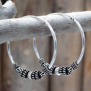 Puede incluir: Un par de pendientes de aro plateados con detalles ornamentados en negro y plata. Los pendientes se muestran en una rama de madera sobre un fondo natural borroso. El diseño presenta una serie de cuentas texturizadas.
