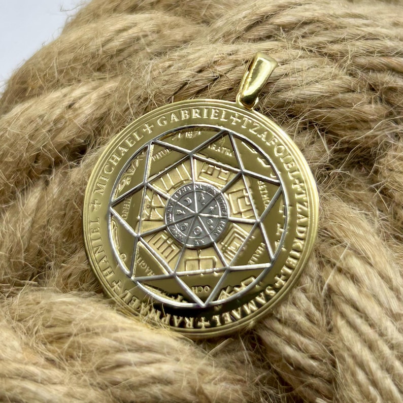May include: A gold-colored pendant with a detailed geometric design and the names of archangels inscribed around the edge. The pendant has a silver-colored center and a gold bail for a chain. The pendant is resting on a brown rope.