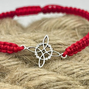 May include: A red macramé bracelet with a silver Celtic knot charm. The bracelet is made of woven red threads and features a detailed silver charm in the centre. The bracelet is displayed on a textured, neutral-toned surface.