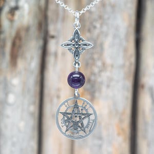 May include: A silver pendant necklace with a cross-shaped top, a purple gemstone, and a pentagram pendant. The pentagram is within a circular frame with Hebrew text. The necklace is suspended from a silver chain.