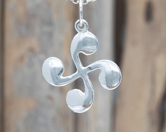 Op de afbeelding: Een gepolijste zilveren hanger in de vorm van een Lauburu, met vier gebogen, komma-achtige armen die vanuit een centraal punt uitstralen. De hanger hangt aan een fijne zilveren ketting met kleine, ronde schakels en een gladde, glanzende afwerking.