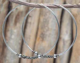 May include: A pair of silver hoop earrings with decorative beaded accents. The earrings are displayed against a natural wood background, showcasing their simple yet elegant design. The hoops are thin and circular, with detailed embellishments.