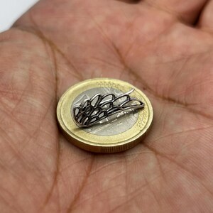 May include: A small, silver-colored wing-shaped charm resting on a Euro coin. The charm has a detailed, openwork design. The coin is gold and silver in color, and the charm is placed on top of it.