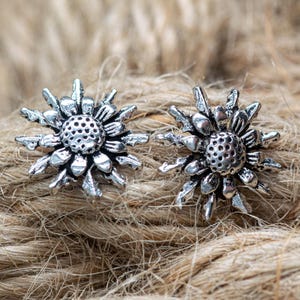 May include: A pair of silver-toned stud earrings. Each earring features a detailed flower design with a central, textured disc surrounded by multiple petals. The earrings are displayed on a textured, brown surface.
