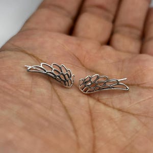 May include: A pair of silver-colored wing-shaped earrings. The earrings are designed with an open, detailed pattern, resembling feathers. The earrings are displayed on a skin-toned background.