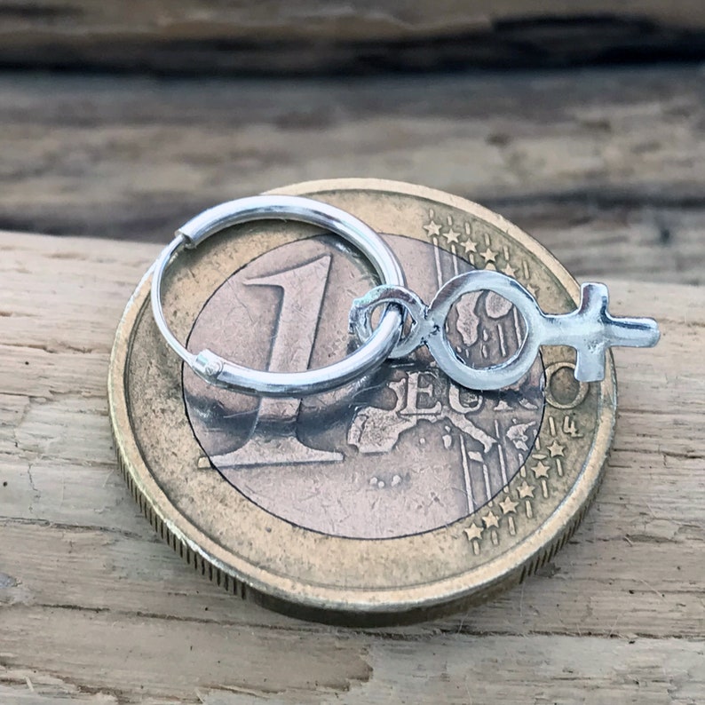 以下が含まれることがあります： 銀製のフープピアスに、女性のシンボルを模した小さな銀製のチャームが付いています。ピアスはユーロ硬貨の上に置かれています。