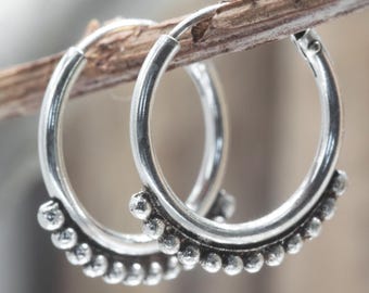 May include: Silver hoop earrings with a beaded design along the lower edge. The earrings are circular and have a simple, elegant design. They are displayed on a brown twig against a blurred background.