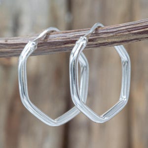 May include: A pair of silver-colored, geometric hoop earrings. The earrings have a unique, angular design, with a hexagonal shape. The earrings are displayed on a thin, brown twig against a blurred wooden background.