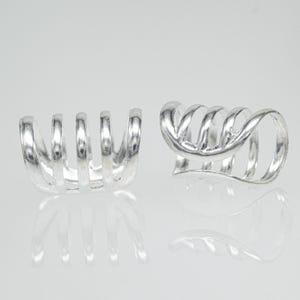 May include: Two silver ear cuffs. One features vertical bars, the other a spiral design. The jewellery is displayed on a reflective surface, creating a mirrored effect. The cuffs are made of a shiny metal.