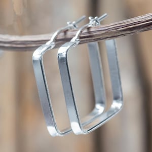 Può includere: Un paio di orecchini a cerchio quadrati argentati. Gli orecchini sono appesi a un sottile ramo marrone. I cerchi hanno una superficie lucida e riflettente e un design semplice e moderno. Lo sfondo è un morbido mix sfocato di marrone e beige.