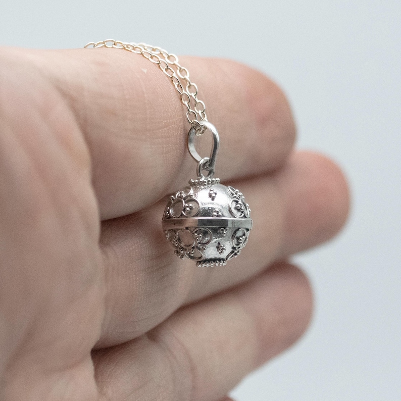 May include: A silver necklace with a delicate, ornate, round pendant. The pendant has a detailed design and appears to be a bell or chime.