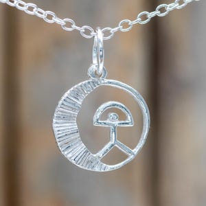 Könnte beinhalten: Eine silberne Halskette mit Anhänger. Der Anhänger hat ein kreisförmiges Design mit einem Halbmond und einer stilisierten Figur. Er hängt an einer filigranen Silberkette. Das Design ist minimalistisch und modern, mit klaren Linien und geometrischen Formen.