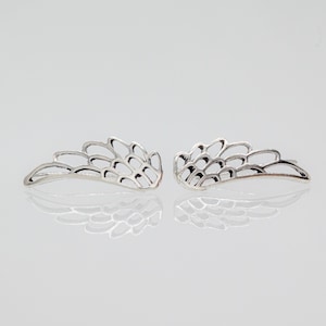 May include: A pair of silver-colored wing-shaped earrings. Each earring features a detailed, openwork design with curved lines and a feather-like appearance. The earrings are displayed on a reflective surface, enhancing their metallic sheen.