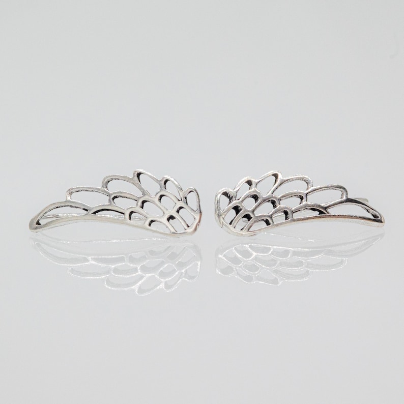 May include: A pair of silver-colored wing-shaped earrings. Each earring features a detailed, openwork design with curved lines and a feather-like appearance. The earrings are displayed on a reflective surface, enhancing their metallic sheen.