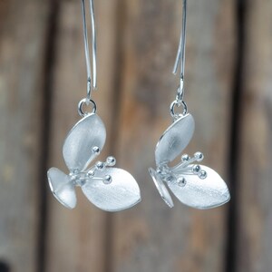 Pode incluir: Um par de brincos pendurados prateados. Cada brinco apresenta um design floral com três elementos em forma de pétala e pequenos detalhes redondos. Os brincos pendem de fios finos e curvos, mostrando um estilo delicado e elegante.