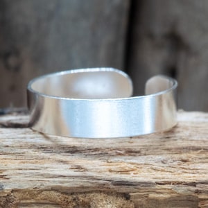 May include: A silver-colored adjustable ring with a flat band. The ring is open at the top and sits on a piece of weathered wood. The ring has a brushed metal finish.
