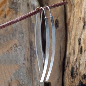 May include: A pair of silver-colored, linear drop earrings. The earrings are suspended from a thin wire and feature a long, flat, rectangular design. The earrings are hanging from a brown twig against a blurred wooden background.