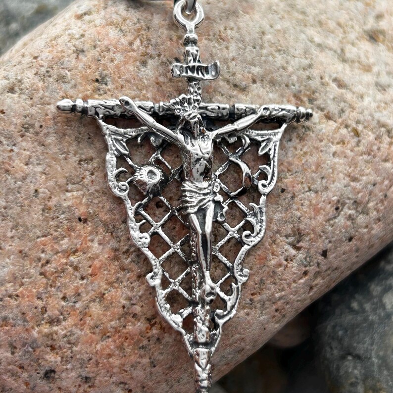 May include: A silver-colored crucifix pendant with a detailed depiction of Jesus Christ on the cross. The pendant features intricate filigree and a decorative banner design. The cross is suspended from a chain.