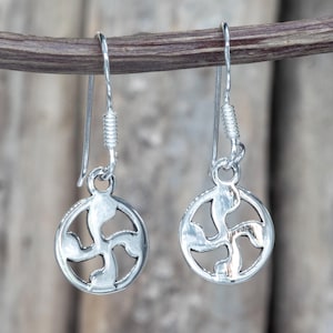 May include: A pair of silver dangle earrings. Each earring features a circular pendant with a stylized, openwork design. The earrings hang from a simple hook with a coiled accent. The background is blurred, suggesting an outdoor setting.