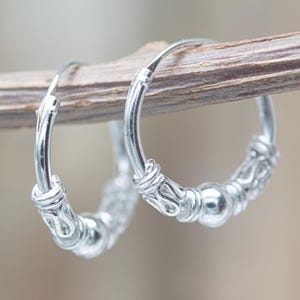 Op de afbeelding: Een paar zilveren oorringen met een gepolijste afwerking. Elke ring is versierd met een centrale gladde bolvormige kraal, geflankeerd door twee getextureerde, decoratieve cilindrische accenten, wat zorgt voor een gedetailleerd metalen ontwerp.