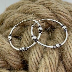 May include: A pair of silver hoop earrings featuring a smooth circular band accented with three evenly spaced polished silver beads, each flanked by small decorative twisted wire rings that provide a textured contrast to the metal surface.