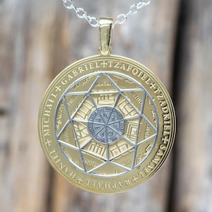 May include: A gold-colored pendant with a silver-colored geometric design and etched text. The pendant hangs from a silver chain. The design features a central circle surrounded by a star-like pattern and inscribed text.