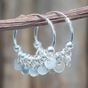 Peut inclure: Boucles d'oreilles créoles argentées ornées de petits disques pendants. Les boucles sont présentées sur une brindille brune, avec un fond flou. Chaque boucle a des perles argentées et une fermeture à cliquet.