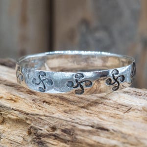 Peut inclure: Une bague en argent ornée d'un motif répétitif de motifs symétriques stylisés. La bague est posée sur un fond en bois naturel, soulignant son aspect artisanal. Le design de la bague est un bijou unique et accrocheur.
