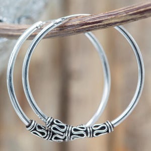 Puede incluir: Un par de pendientes de aro de plata con una banda circular lisa y pulida. Cada pendiente está decorado con una cuenta de plata texturizada en la parte inferior, que muestra un patrón intrincado y repetitivo de ondas con detalles oscurecidos.