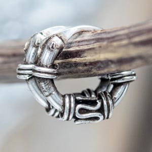 May include: A silver-colored ring with an intricate design, featuring a series of connected loops and decorative elements. The ring is wrapped around a brown, textured object, possibly a piece of wood or antler. The ring's design suggests a vintage or handcrafted style.
