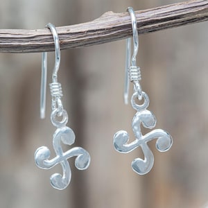 May include: A pair of silver-toned drop earrings. Each earring features a stylized, swirling design suspended from a hook. The earrings are displayed against a blurred, natural background, highlighting their intricate details and reflective surface.