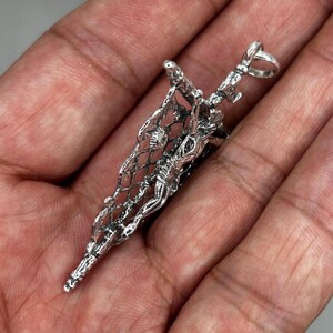 May include: A detailed silver pendant featuring a figure within a net-like structure. The pendant has intricate detailing and a loop at the top for a chain. The design is highly textured and appears to be handcrafted, showcasing a unique artistic style.
