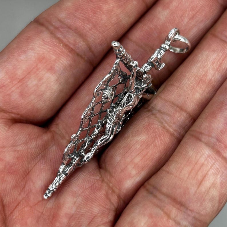 May include: A detailed silver pendant featuring a figure within a net-like structure. The pendant has intricate detailing and a loop at the top for a chain. The design is highly textured and appears to be handcrafted, showcasing a unique artistic style.