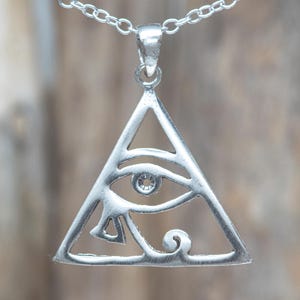 Puede incluir: Un colgante plateado con forma de triángulo que contiene el símbolo del Ojo de Horus. El colgante cuelga de una cadena plateada sencilla. El diseño presenta detalles recortados que forman el ojo estilizado, la pupila y líneas decorativas.