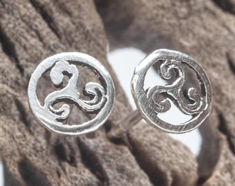May include: A pair of silver-colored stud earrings. Each earring features a circular design with a Celtic triskele symbol. The earrings are displayed on a textured, brown surface.