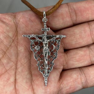 May include: Silver-toned pendant necklace featuring a detailed crucifix design. The pendant is suspended from a brown leather cord. The crucifix depicts Jesus on the cross, with intricate filigree detailing around the edges. The pendant is approximately 2 inches in length.