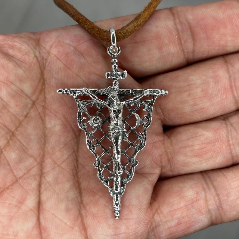 May include: Silver-toned pendant necklace featuring a detailed crucifix design. The pendant is suspended from a brown leather cord. The crucifix depicts Jesus on the cross, with intricate filigree detailing around the edges. The pendant is approximately 2 inches in length.