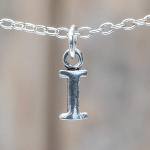 Op de afbeelding: Zilverkleurige ketting met een hanger in de vorm van de letter "I". De hanger is een gedetailleerd, driedimensionaal ontwerp. De ketting is gemaakt van kleine, ovale schakels.